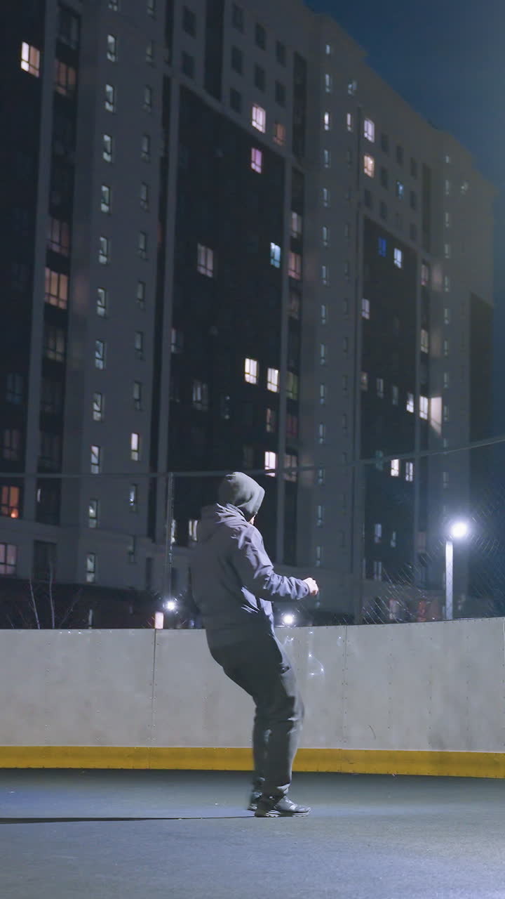 Back view of man in hoodie playing soccer against wall under bright stadium lighting at night, soccer ball near wall reflects silhouette with urban cityscape and illuminated apartment buildings