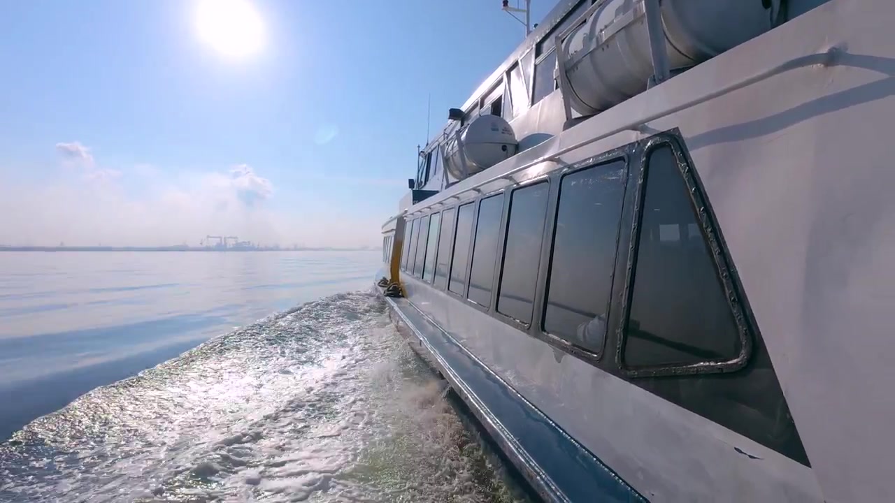 Eine Aufnahme aus der Ich-Perspektive vom Deck einer fahrenden Fähre, die eine weiße Kielwelle und eine ferne Stadtsilhouette entlang einer wichtigen Schifffahrtsroute in Südostasien oder der Straße von Singapur zeigt.