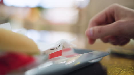 blurry view of tray with kfc bag on table and hand dipping inside to pick chips, soft focus suggesting casual meal setting, light reflections on surface, background indistinct, fast food atmosphere