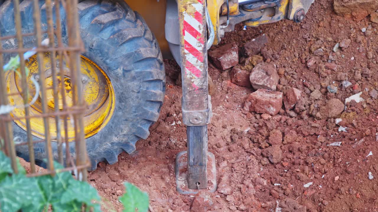 Primer plano del brazo hidráulico de una excavadora o retroexcavadora y una rueda grande, trabajando en una obra en construcción. De día, toma estable, 4K