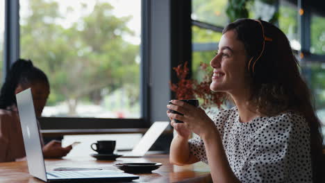 businesswoman wearing wireless headphones working on laptop in cafe shop with colleague in background