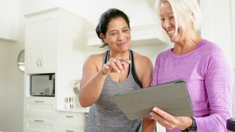 Dos-Mujeres-Mayores-Felices-Y-Diversas-Usando-Tableta-Y-Riéndose-En-La-Cocina,-Cámara-Lenta