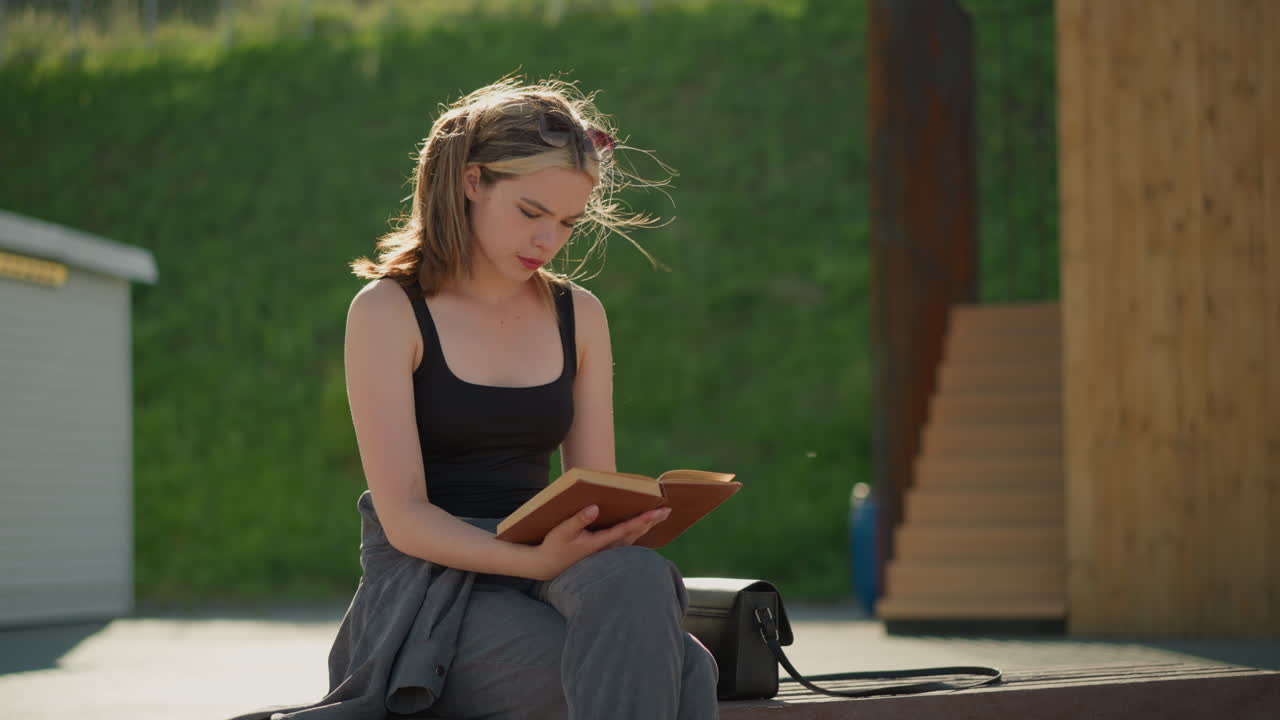 Lady in seated with legs crossed reading a book. Wind blows her hair while her hand rests on the book, a black bag is next to her, with a blurred background featuring greenery and wood structures