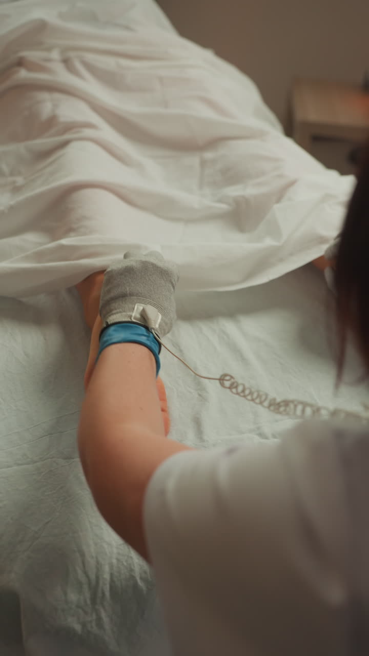 Terapista applica un elettrodo a guanto conduttivo lungo polpaccio e caviglia sotto un lenzuolo, connettore a catena sottile visibile, illuminazione clinica, movimento mirato per il sollievo dal dolore e il miglioramento della circolazione, professionista.