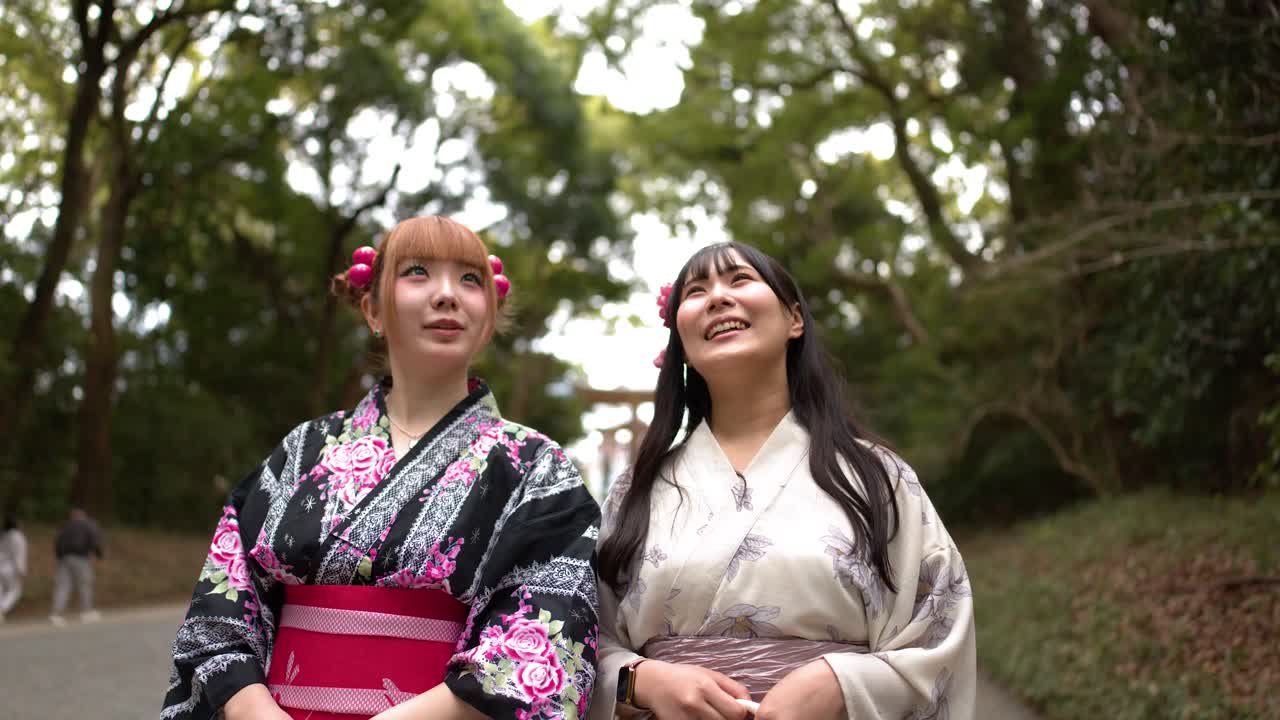 Duas amigas em quimonos florais caminham e conversam por uma exuberante trilha verde em parque florestal.
