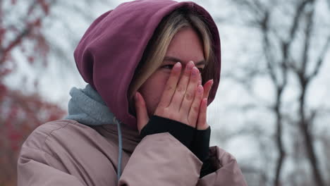 girl in winter outfit standing in cold, blowing hot air into hands to stay warm, hooded jacket and layered clothing emphasize the freezing weather, blurred background of trees