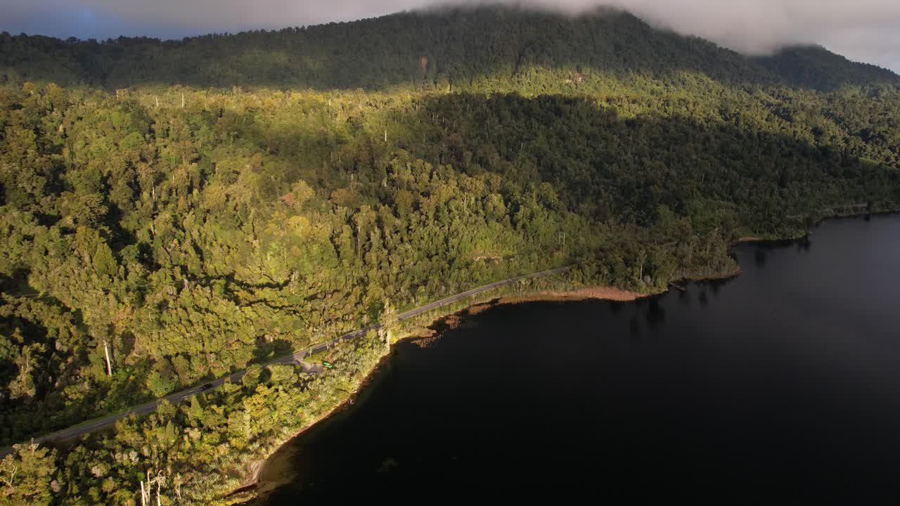 Winding Road Skirting Calm Dark Lake Bordered By Dense Forest With Sunlight. Lake Ianthe In New Zealand. aerial shot