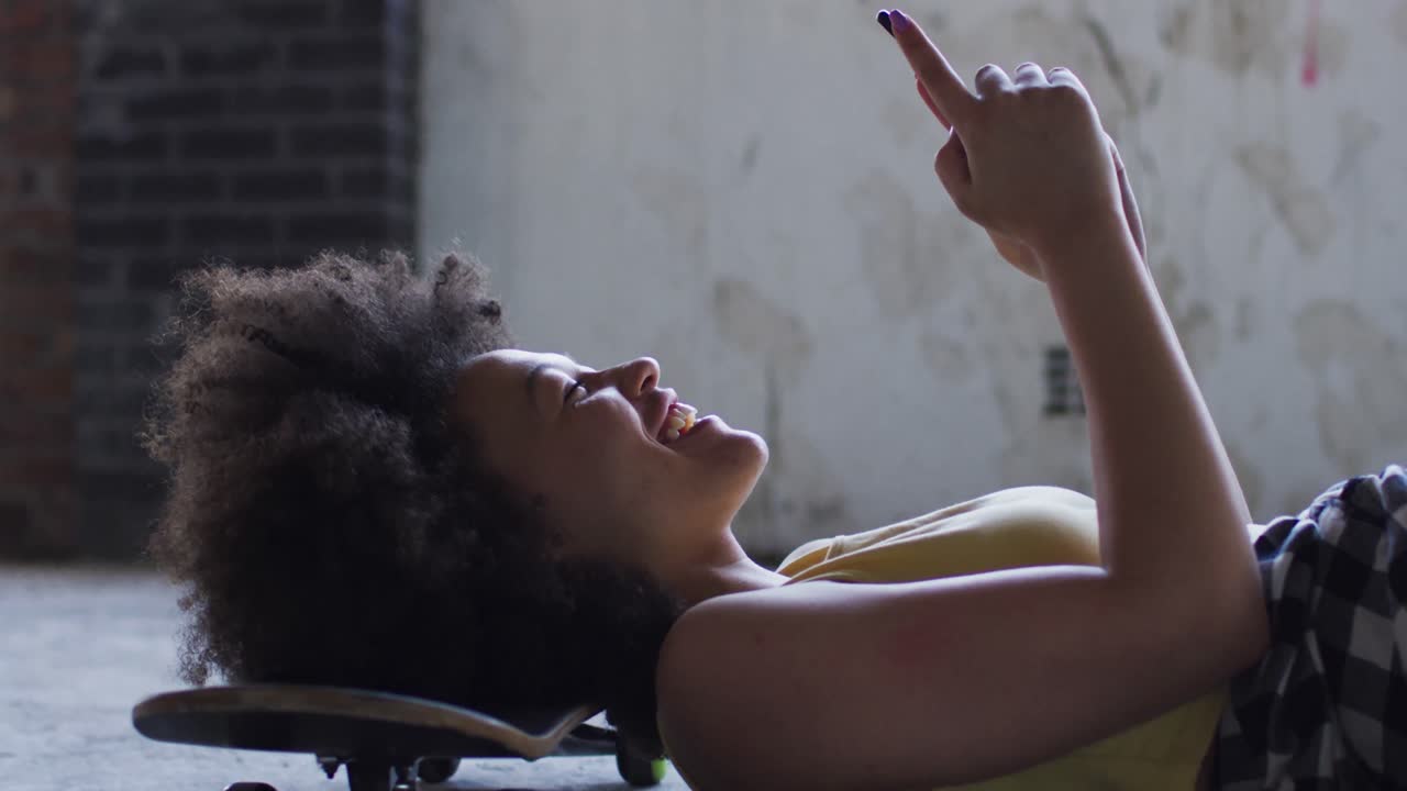 Mixed race woman in an empty building lying on a skateboard using smartphone laughing