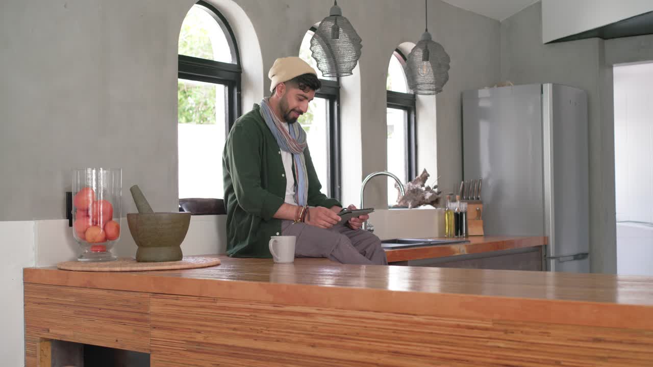 Homem indiano sentado em ilha de cozinha de madeira, após usar o tablet, pegando uma caneca branca para beber