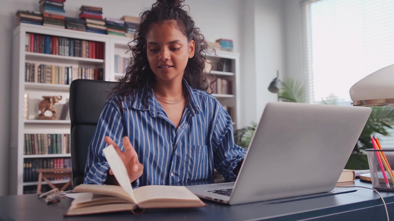 Millennial indian girl sit at desk in living room study on laptop making notes