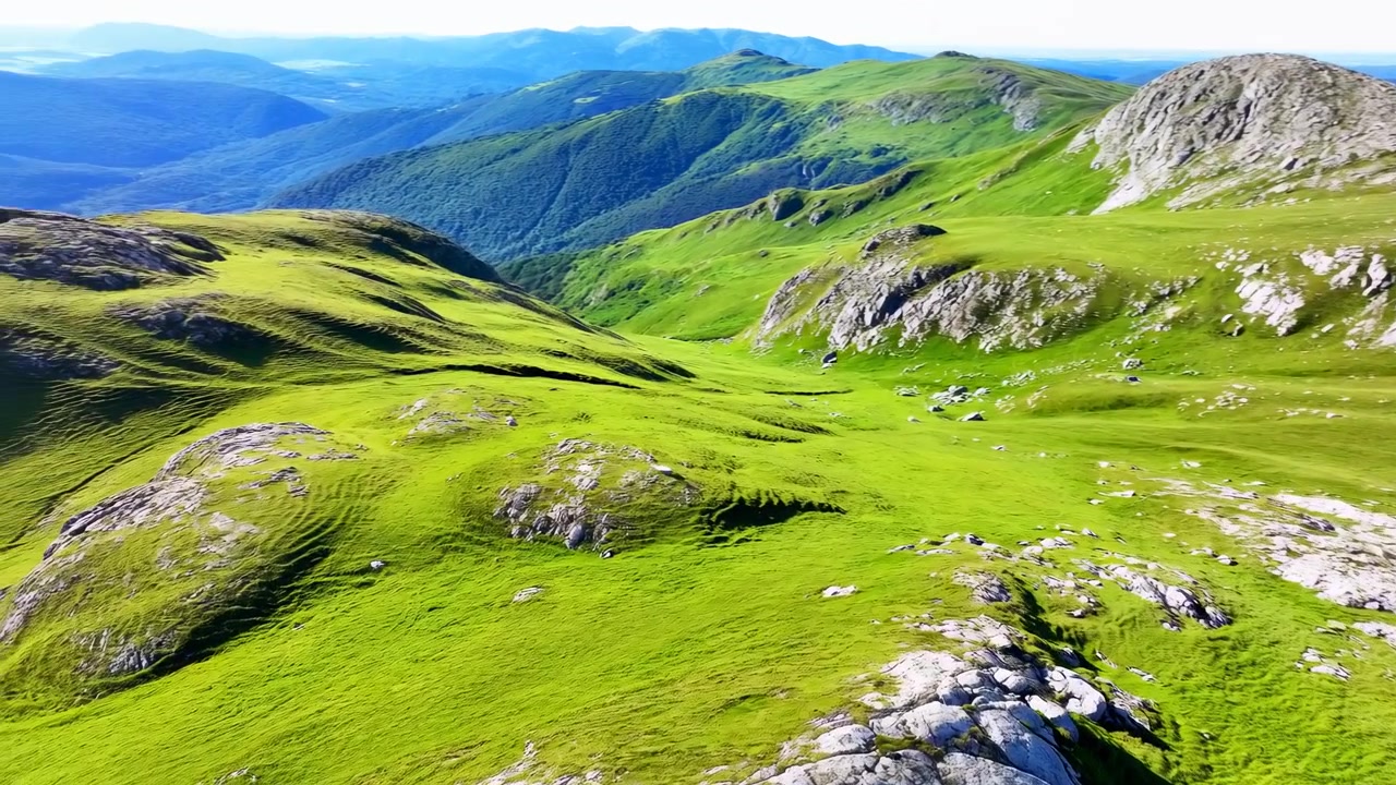 Vista obliqua elevata su un vasto paesaggio montano con colline ondulate coperte d'erba, altopiano roccioso, roccia affiorante, massi chiari sparsi, vetta distante, cielo azzurro limpido e mezzogiorno luminoso.