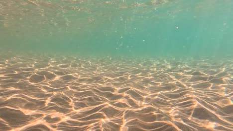 clear underwater view of a sandy sea floor with sunlight patterns, gentle water movement, and turquoise tones. natural lighting, static camera, tranquil mood
