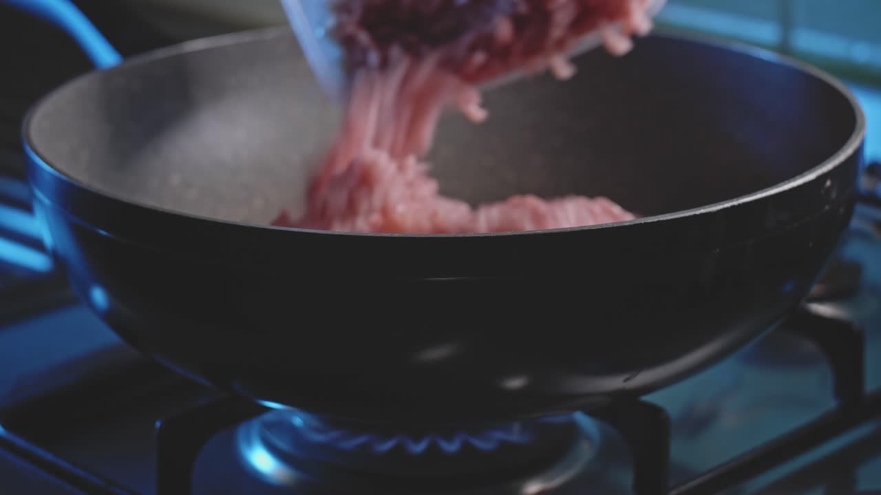Side view close up of raw ground or minced meat getting added into a deep nonstick frying skillet pan heating up on a clean blue flame from a builtin hob cooktop gas stove