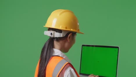 close up back view of asian female engineer with safety helmet working on a green screen laptop and looking around while standing in the green screen background studio