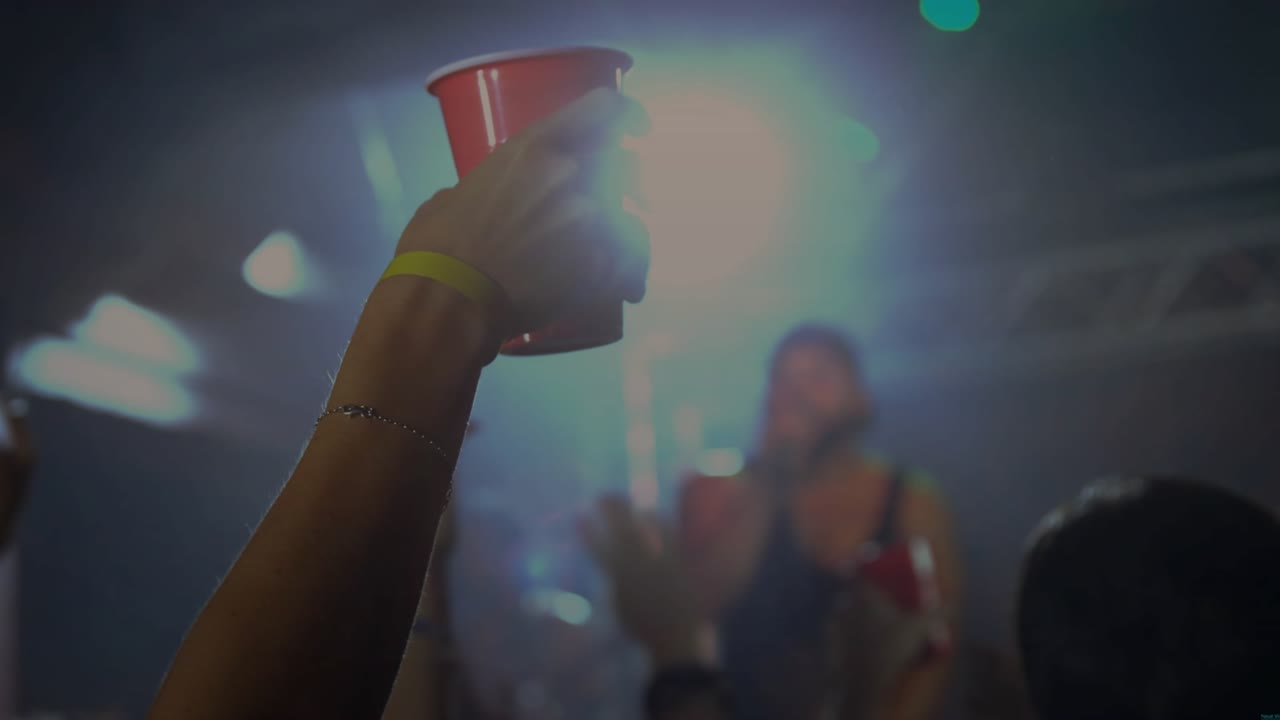 Performer lifting cup, arm holding red cup as crowd raising cups, lights haloing crowd at concert