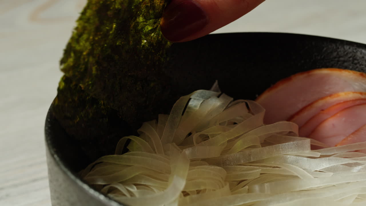 Woman adding Nori to a Bowl of Noodles with Ham