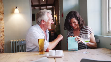 senior white man giving a present in a gift bag to his hispanic female friend, sitting with drinks at a table in a pub, close up, side view
