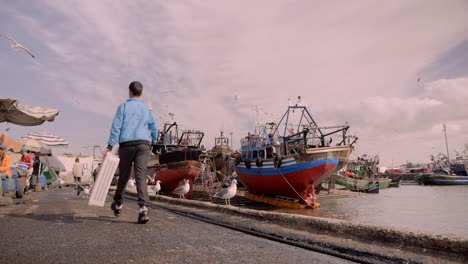 wide establishing shot, man walks with plastic crate through port city of essaouira morocco