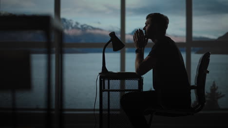 The-silhouette-of-a-thoughtful-man-sitting-at-a-table-against-the-background-of-a-window-with-oceans-and-the-sea.-A-restless-man-sitting-at-a-table-with-a-desk-lamp