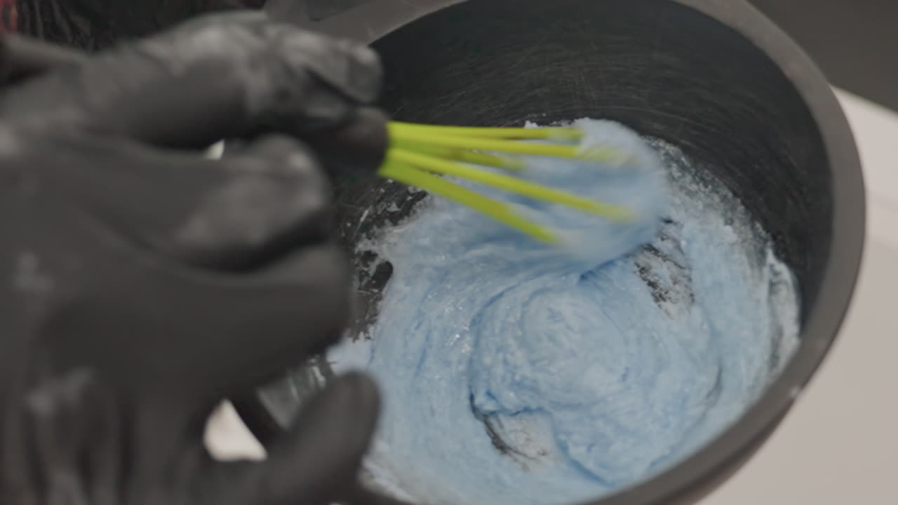 Unos guantes negros mezclan un cuenco con polvo azul; una técnica de estética clínica mezcla pigmento en polvo hasta obtener una pasta homogénea en un recipiente negro; primer plano de unas manos con guantes de nitrilo y un batidor verde; estéril