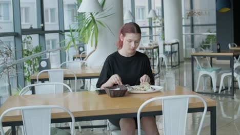 young woman eating a fresh salad at a wooden table in a modern restaurant with large windows, natural light, and urban views. relaxed dining scene