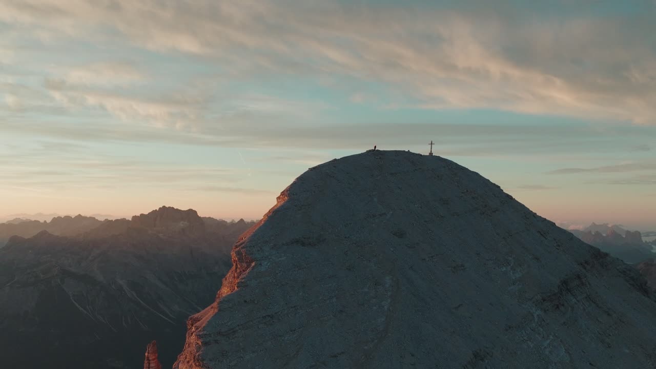 Drone view of a mountaineer on the ridge of Tofana di Rozes in Italy's Dolomites at sunrise, with the summit cross and Monte Antelao visible in the background