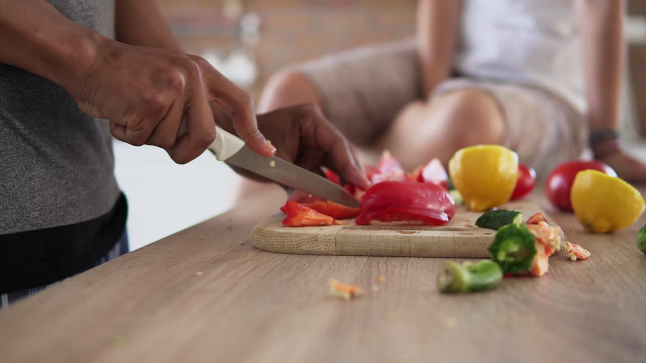 Close Up view of african guy's hands cutting vegetables preparing food while his caucasian girlfriend is sitting on the table in