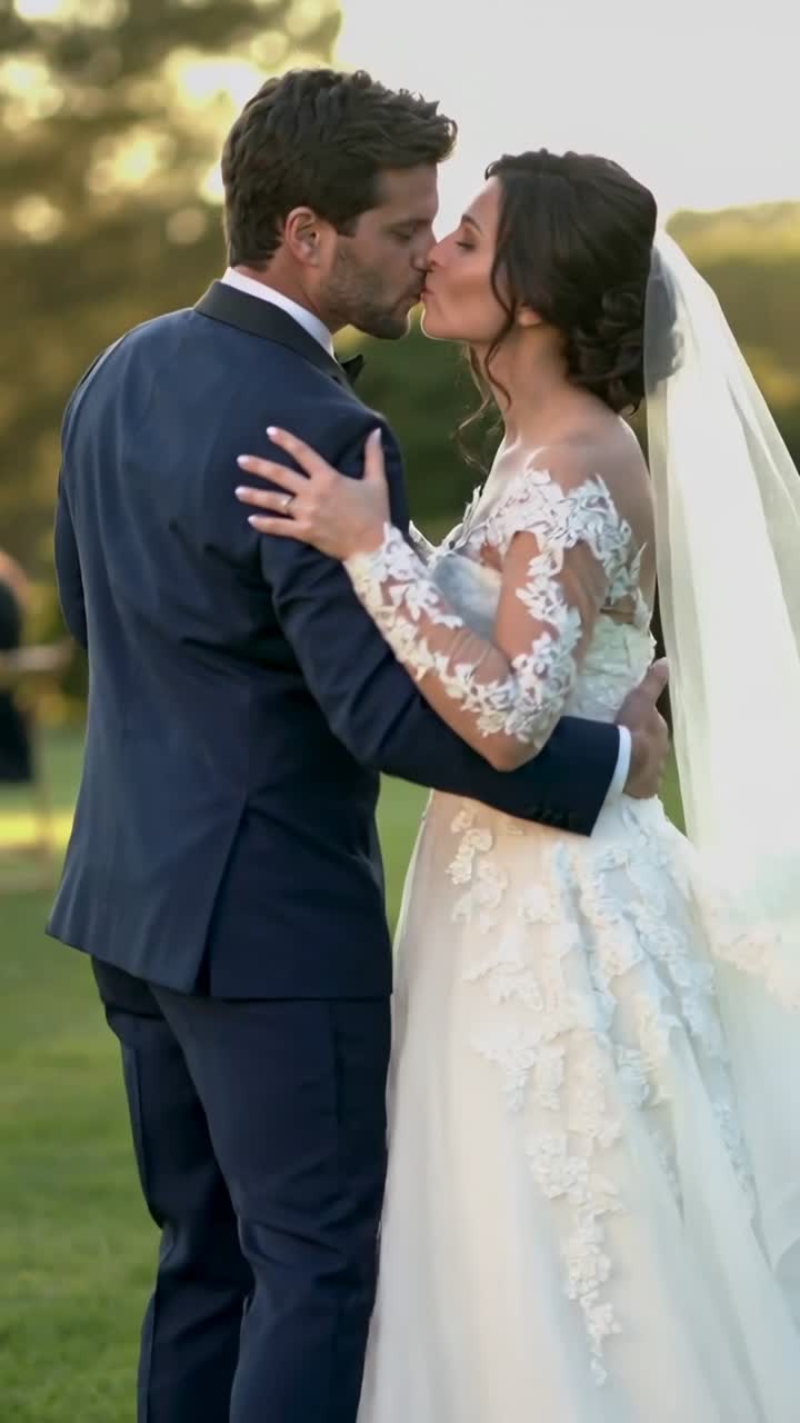 Video vertical: Novio con traje azul marino sosteniendo a la novia con vestido de encaje, inclinándose para besarla en el césped, con el velo de novia.