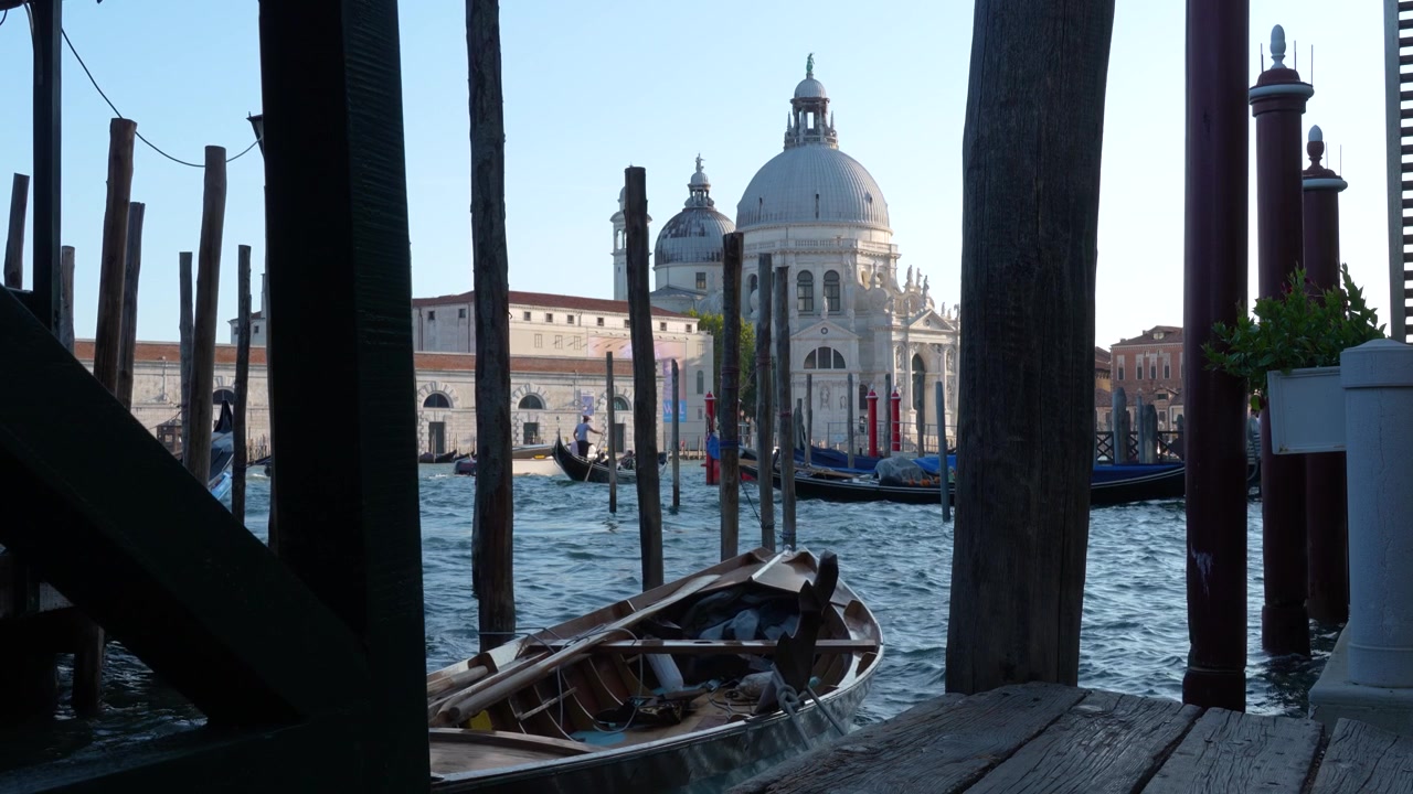 Static cinematic shot of an anchored wooden boat gently moving on the Grand Canal in Venice, with Basilica di Santa Maria della Salute and a distant gondolier in the background