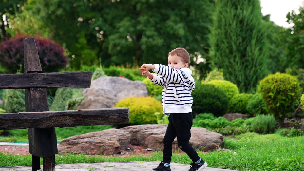 Four-year-old toddler boy runs near the wooden bench. Kid runs in circles holding his hands in front of him.