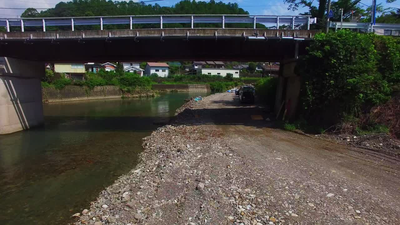 Captivating aerial shot advancing above a river in a serene Japanese village, descending beneath a bridge, displaying abandoned vehicles by the water's edge