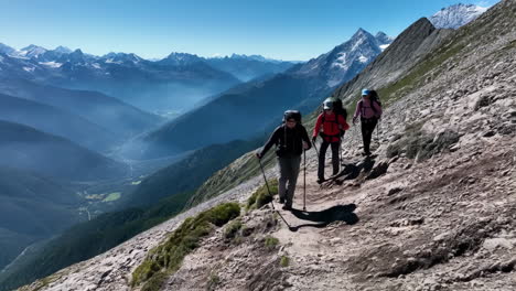 three hikers trekking on a rocky mountain trail with a vast valley and snow-capped peaks in the background