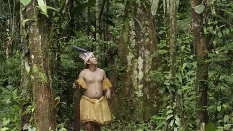 portrait shot of an indigenous guy wearing a feathered hat and fringed shirt walks in the dense forest in leticia, amazon, colombia