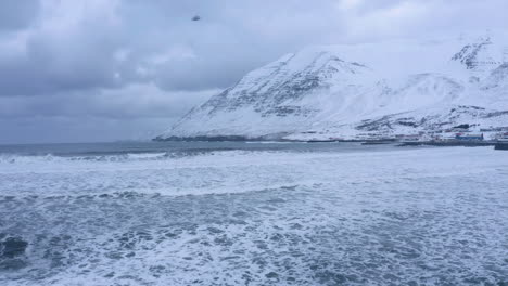 Arctic-ocean-waves-and-winter-in-Olafsfjordur-town,-towering-snow-covered-mountains-in-background--North-iceland-,aerial-flight