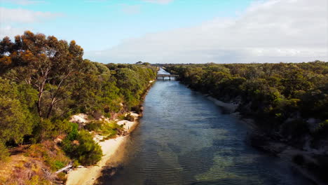 drone view crystal clear river flowing through a small forested valley with a bridge on a partly cloudy day