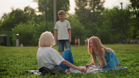 siblings playing and chatting with mom on picnic blanket in sunlit park, girl walking near blanket while brother watches, warm summer light casting soft glow on relaxed family bonding