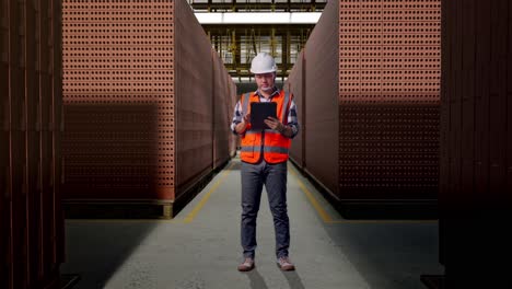 full body of asian male engineer with safety helmet working on a tablet while standing with red brick packed in stacks are stored