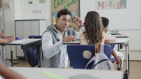 happy diverse male teacher talking to girl at desk in elementary school class, slow motion