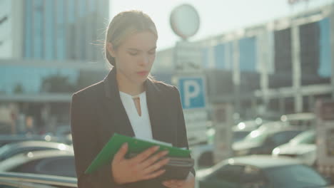 human resource manager in business attire walking outside modern office building reviewing documents in folder under sunlight with parked cars and blurred figures in background