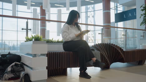 young lady with long dark hair sits on stylish wooden bench in bright modern mall, reading book with focused expression, jacket beside her, surrounded by potted plants, curved bench design