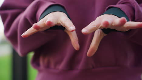 strength trainer in hoodie performs gentle hand stretch by pressing palms together and flexing fingers during calm relaxation before outdoor workout under open sky at urban fitness park