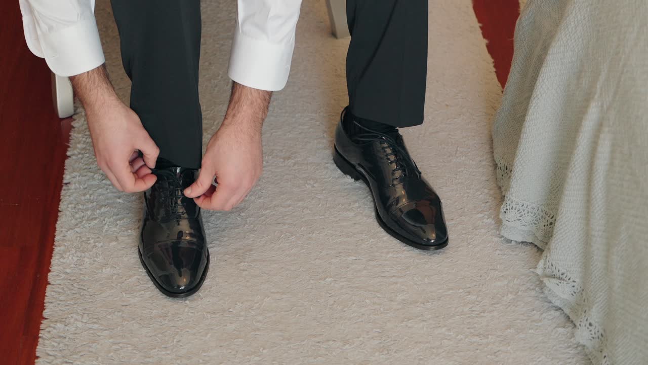close up of man tying black dress shoes on beige carpet during wedding preparation indoors