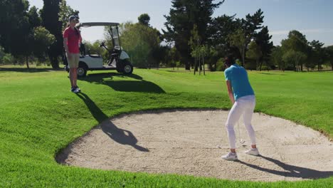 Two-caucasian-women-playing-golf-wearing-face-masks-one-taking-shot-from-bunker