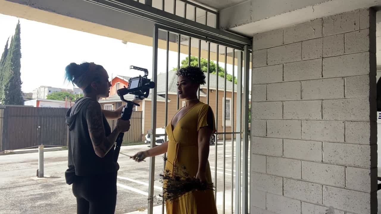 A videographer films a stylish woman in a bright yellow dress next to bars and a white brick backdrop who is holding dried flowers up to the camera