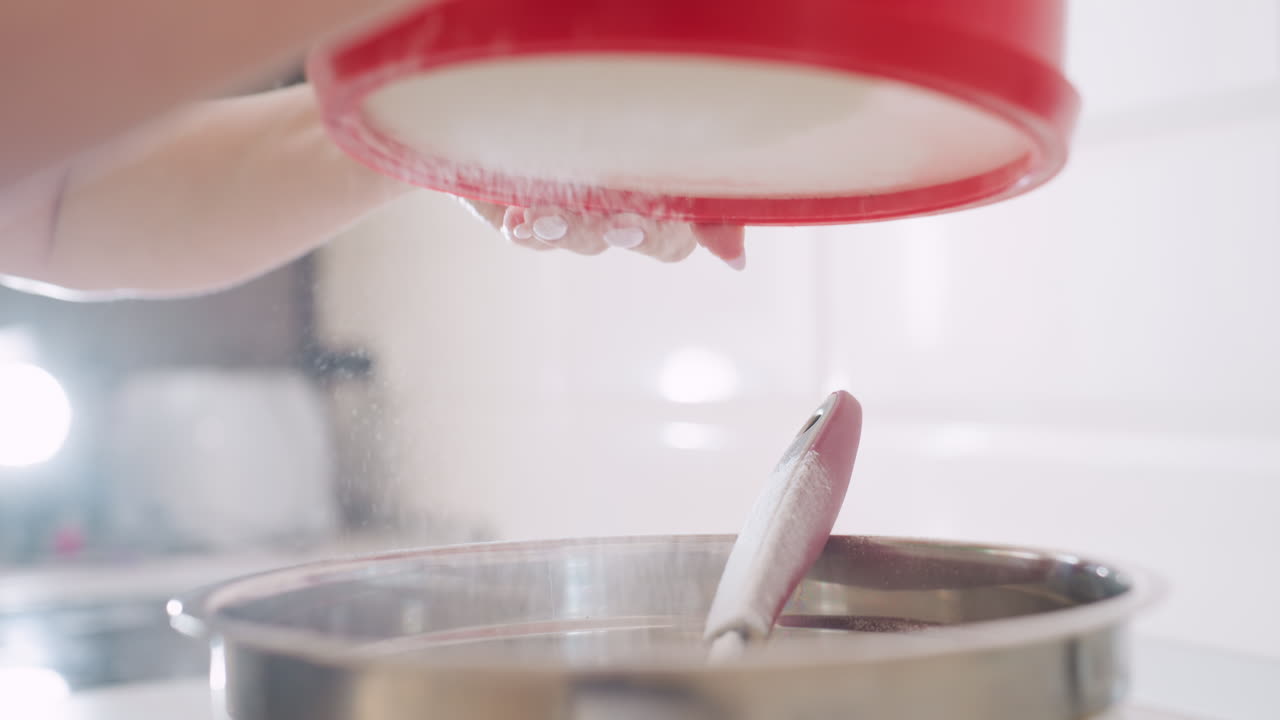 Close up of caterer filtering flour with red sifter into stainless bowl, fine flour dust falling through sieve while mixing utensil rests inside bowl during careful baking preparation in kitchen