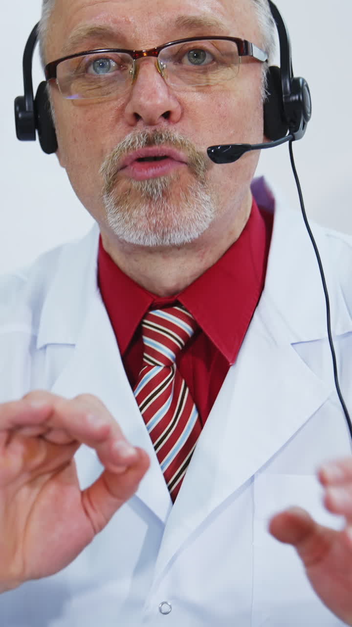 Portrait of senior doctor in glasses and headset. Professional male physician in white medical uniform speaking looking at camera. Vertical video