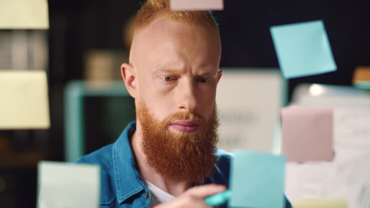 Serious redhead businessman writing on stickers at transparent wall in office.