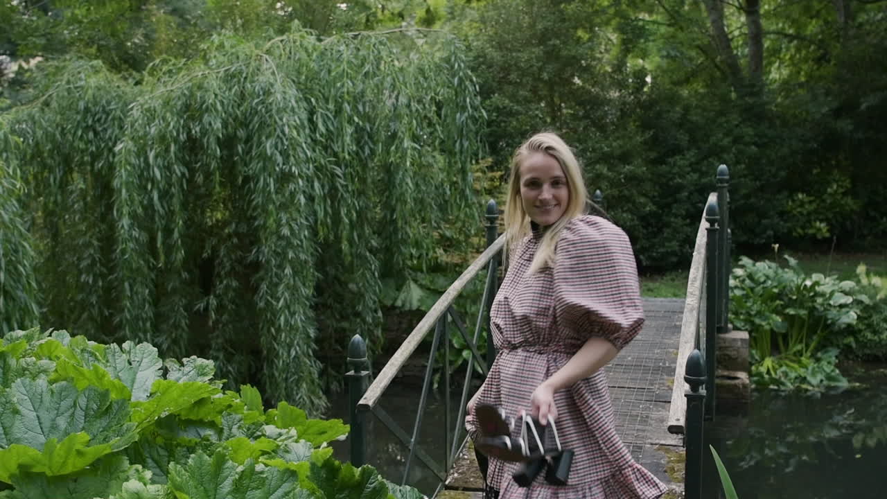 Fashionable woman with blond hair walks proudly over small bridge in garden in slow motion
