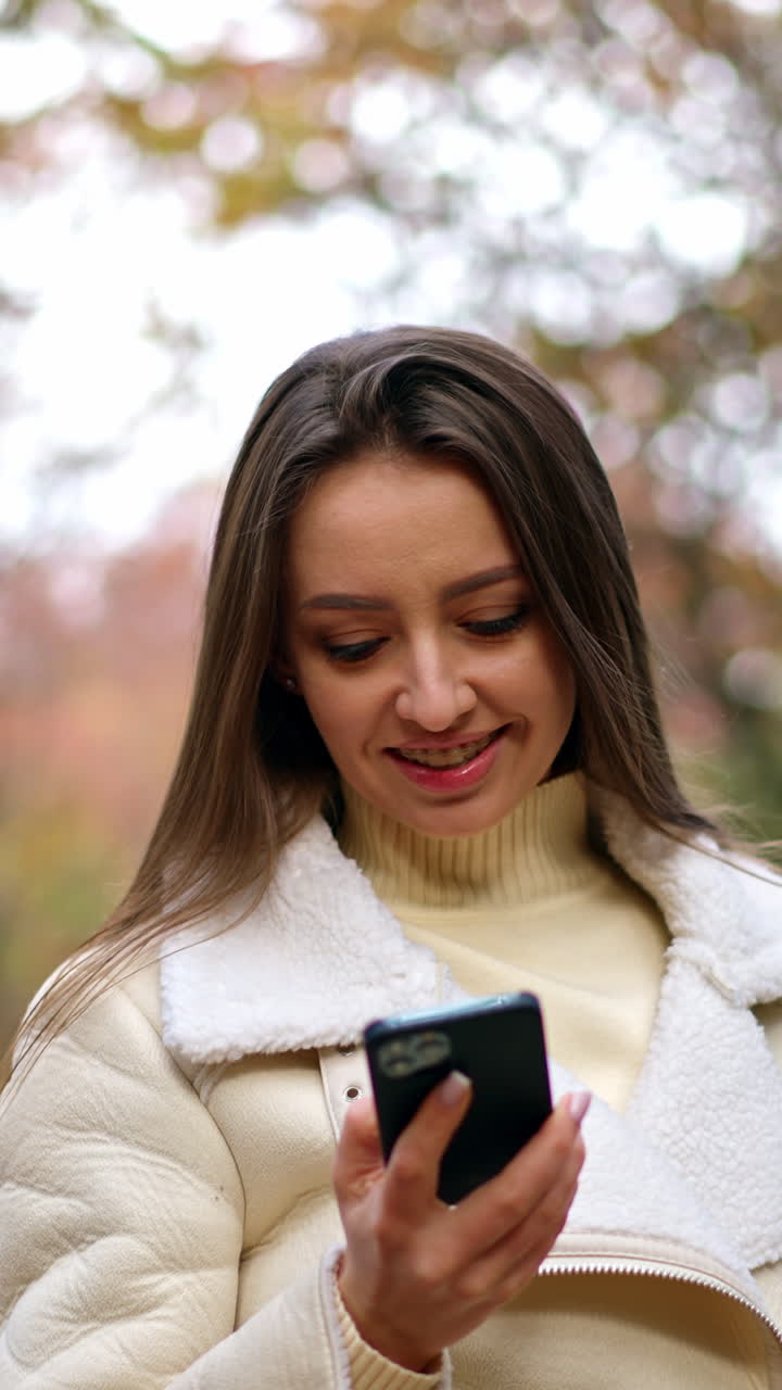Beautiful smiling Caucasian lady speaks on the phone walking by the park. Lady goes looking at phone and then tries to take a picture of autumn trees. Vertical video