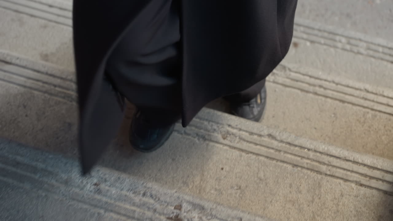 Прочный черный ботинок поднимается по бетонной лестнице, угловой вид подчеркивает канавки для безопасности, потертую текстуру и городскую среду, что предполагает размеренную поездку на работу, минимальную композицию и приглушенный целеустремленный подъем